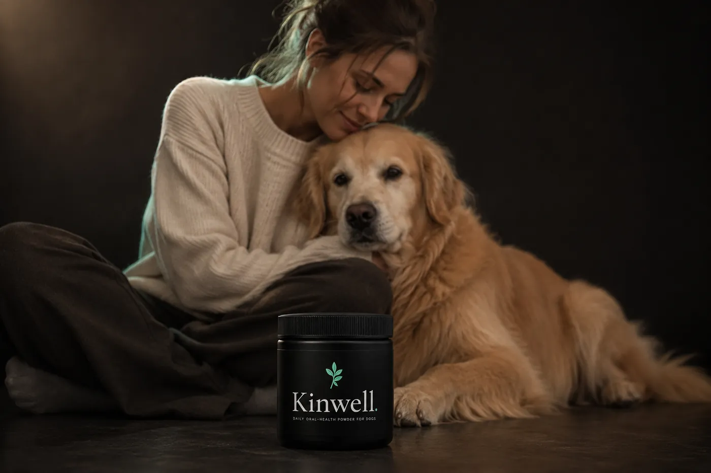 Owner sitting on a dark floor with Bear, her golden retriever, a jar of Kinwell daily oral-health powder in the foreground